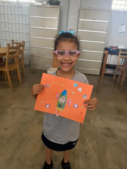 Niña con espejuelos mostrando su cohete sobre papel naranja