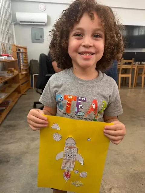 Niño con camiseta STEAM mostrando su cohete espacial en papel amarillo