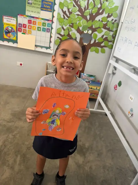 Niña sonriente presentando su dibujo del cohete en papel naranja