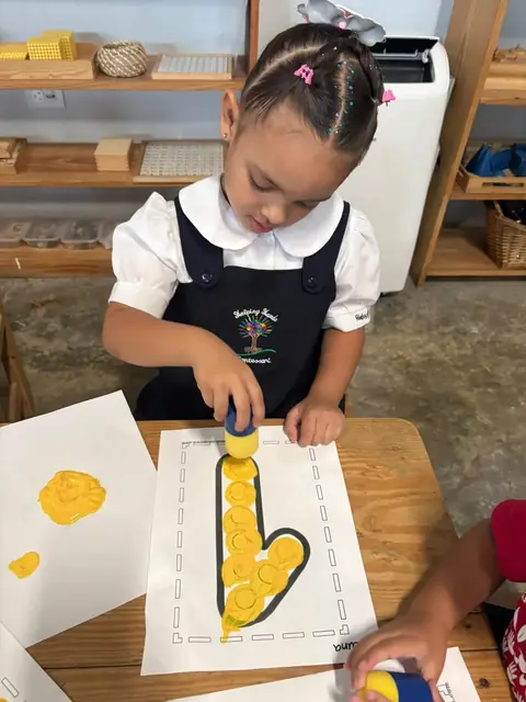 Niña de uniforme trazando su letra con pintura amarilla
