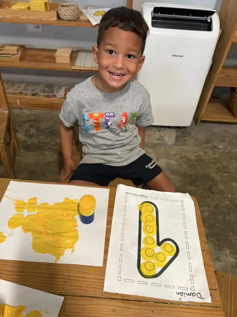 Niño sonriente junto a su hoja de trazos con pintura amarilla