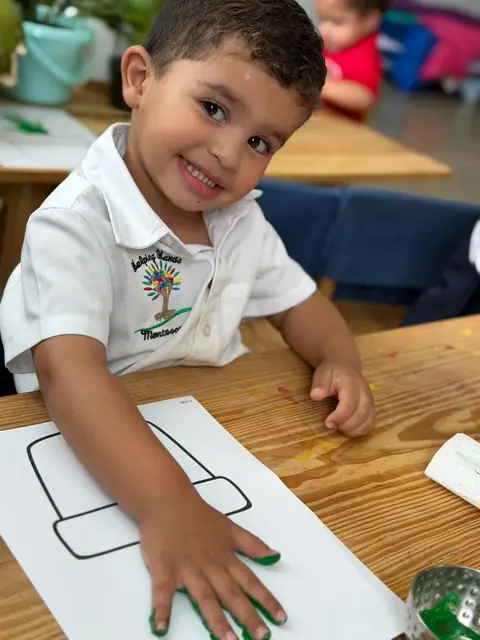 Niño sonriente con los dedos pintados de verde junto a su lámina